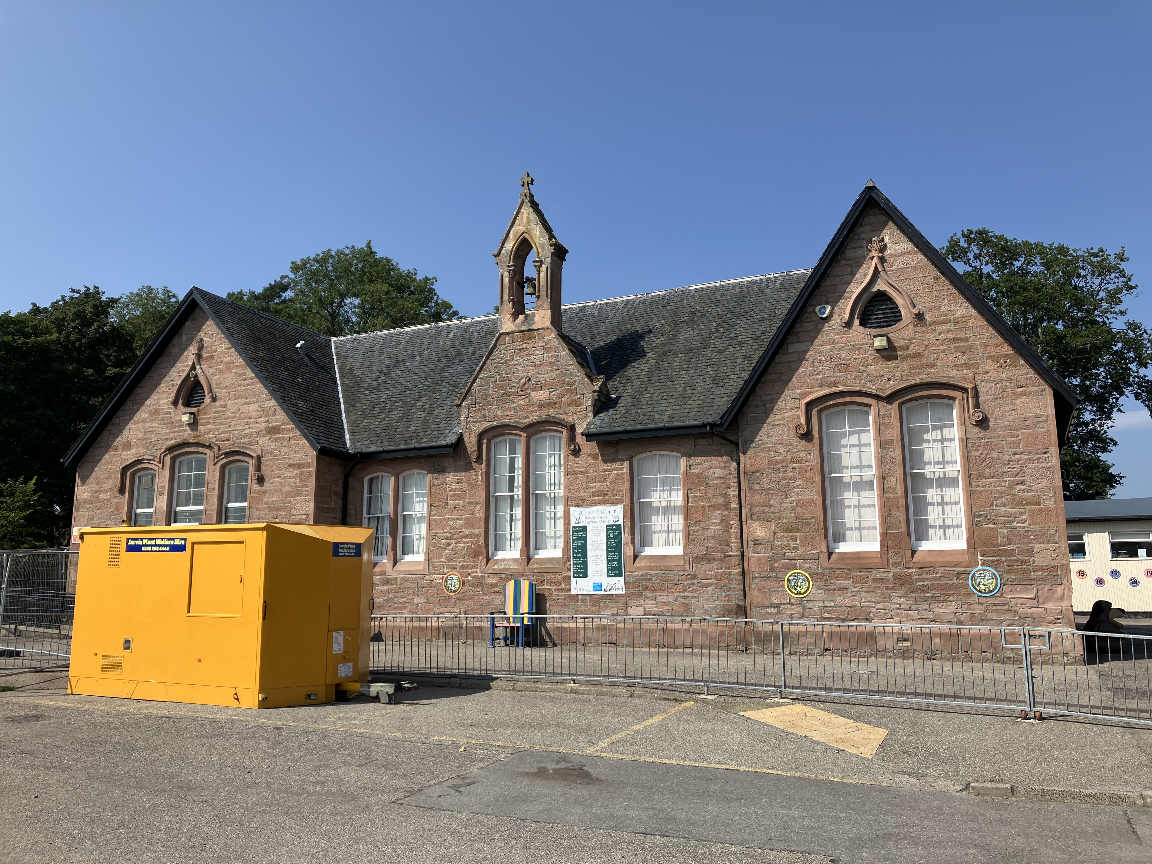 Red sandstone school with central bellcote and yellow container in foreground