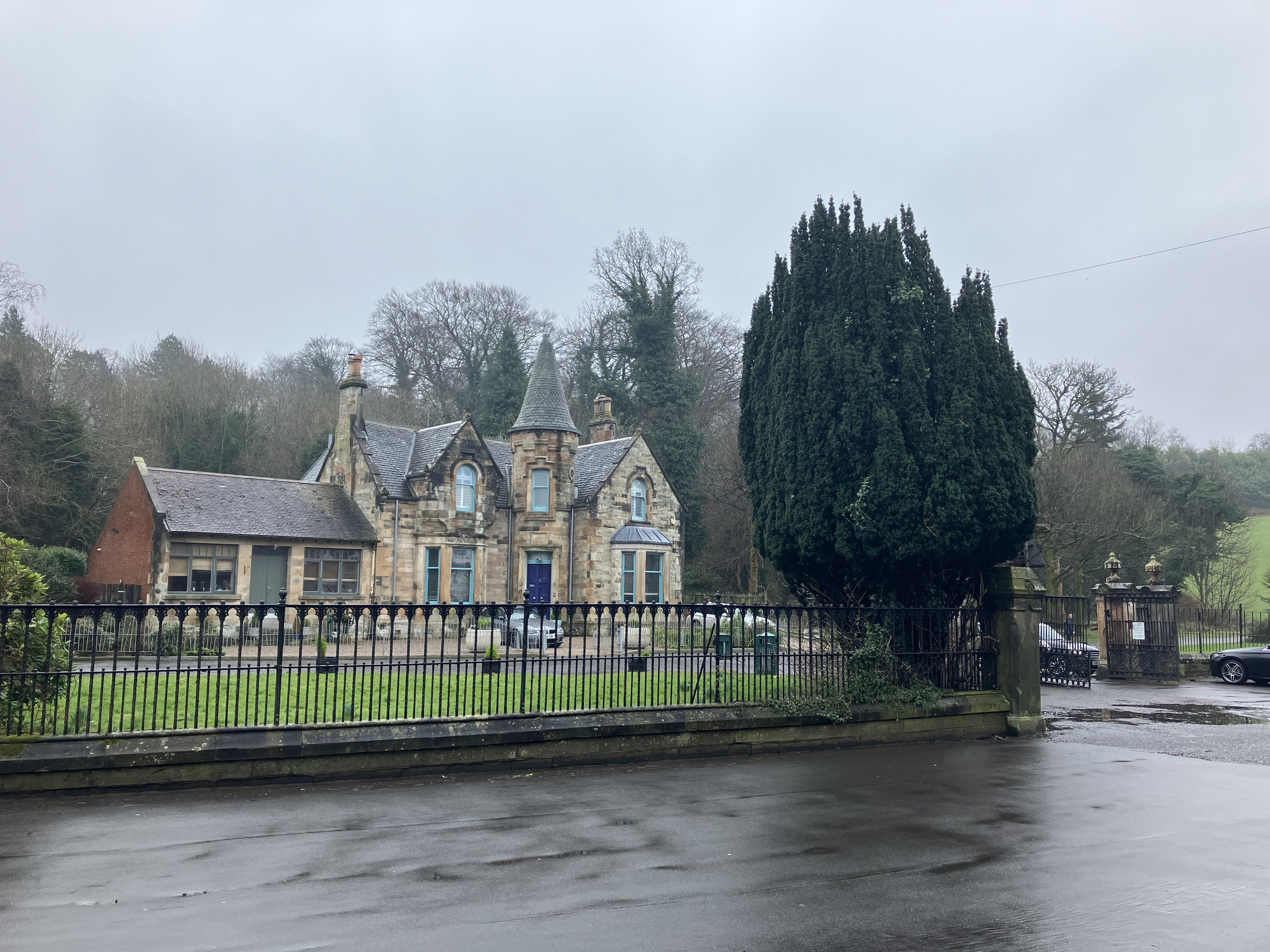 Picture of entrance to Cathcart Cemetary from the street. A black iron fence in the foreground of image border the grounds, with a large green conifer tree beside teh entrance. There are cars in the driveway and a large stone building in the background, surrounded by trees which suggest it is winter time due to the lack of foilage. It is a grey day.