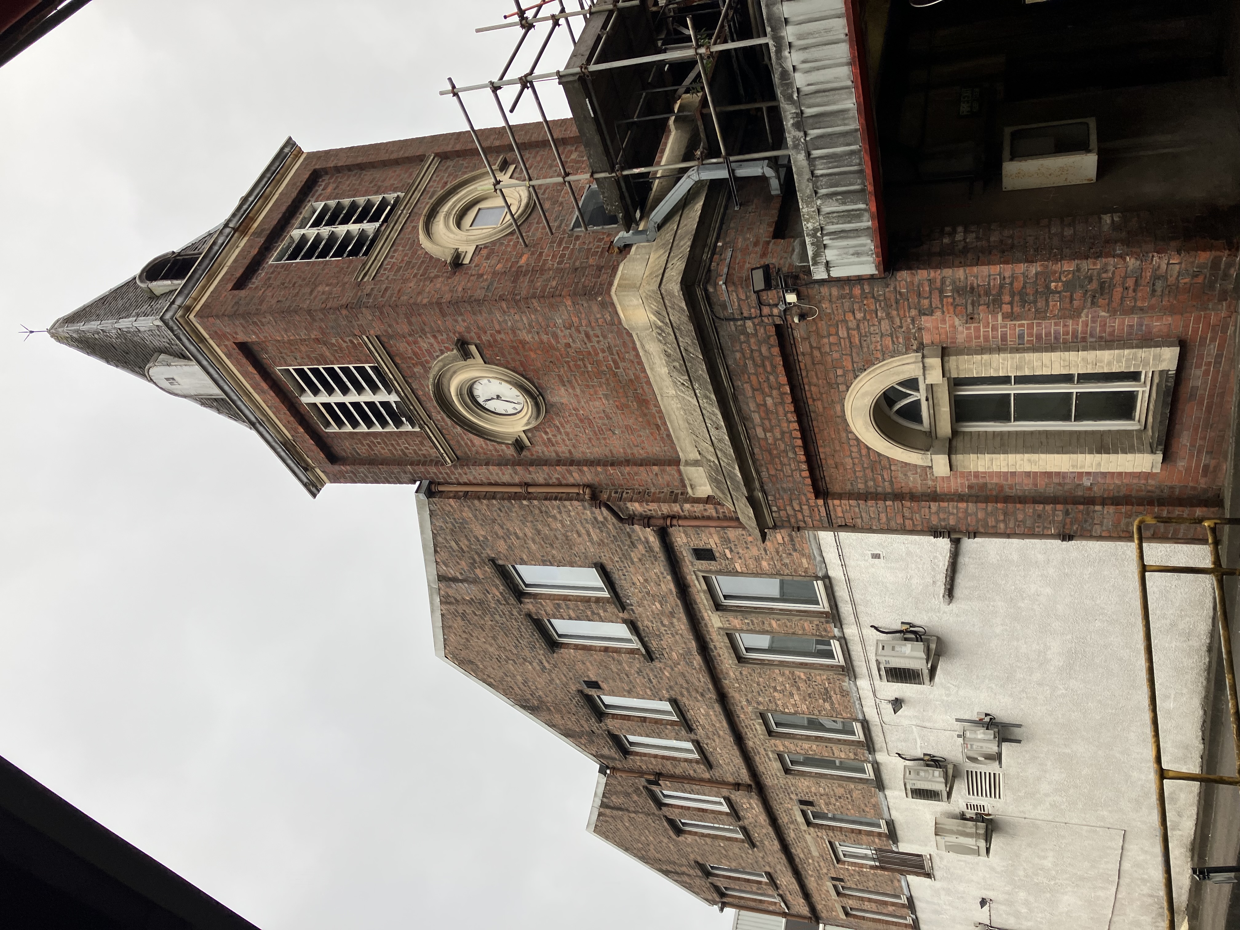 Portrait image of clock tower, which is built with red bricks. Some scaffolding flanks one side of the tower.