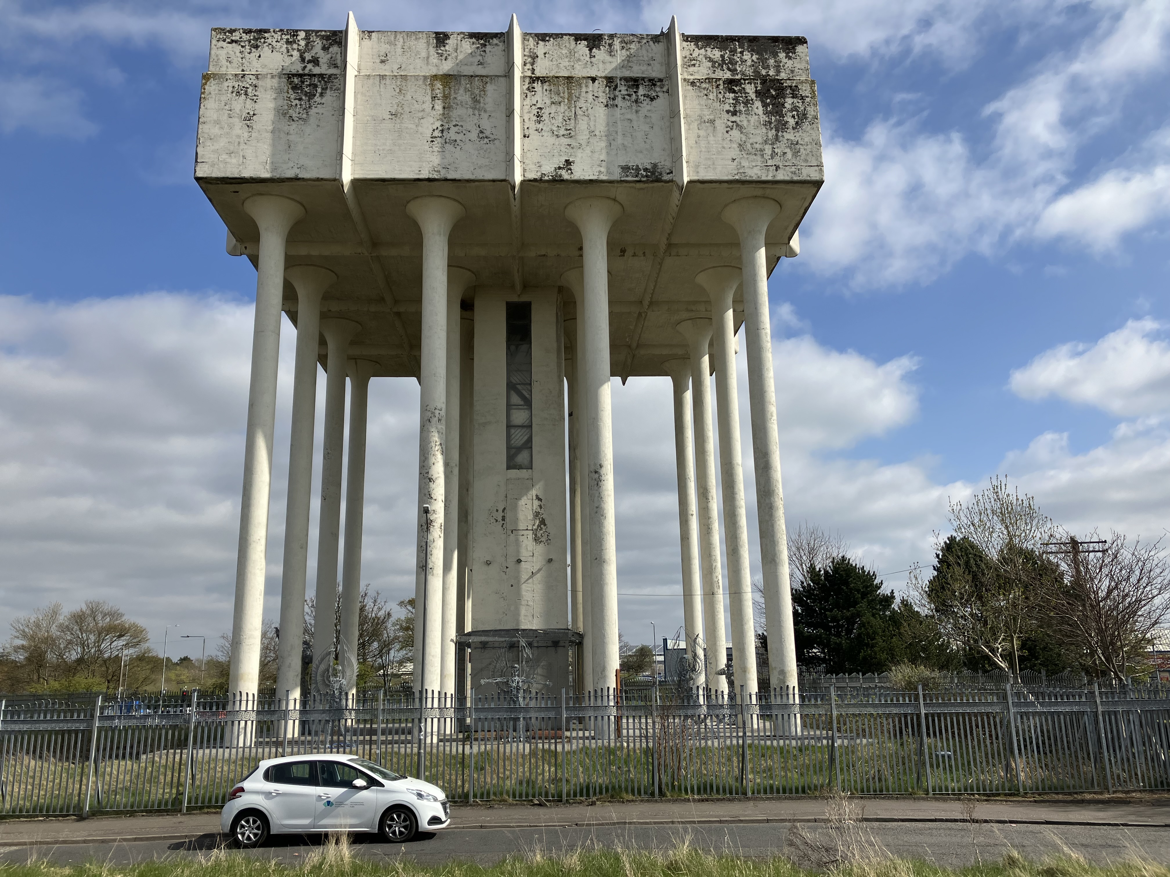 A tall concrete water tower supported by cylindrical pillars under a partly cloudy sky. The structure’s upper tank section appears weathered with visible discoloration. A small white car is parked on the road in front of the fence, with grass and trees in the background.