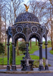 Dalmuir Park Drinking Fountain with Dalmuir Park. From the many leaf-less trees behind the fountain, it appears to be a winter's day. The grass appears lush and the roads around the fountain look wet from recent rain.