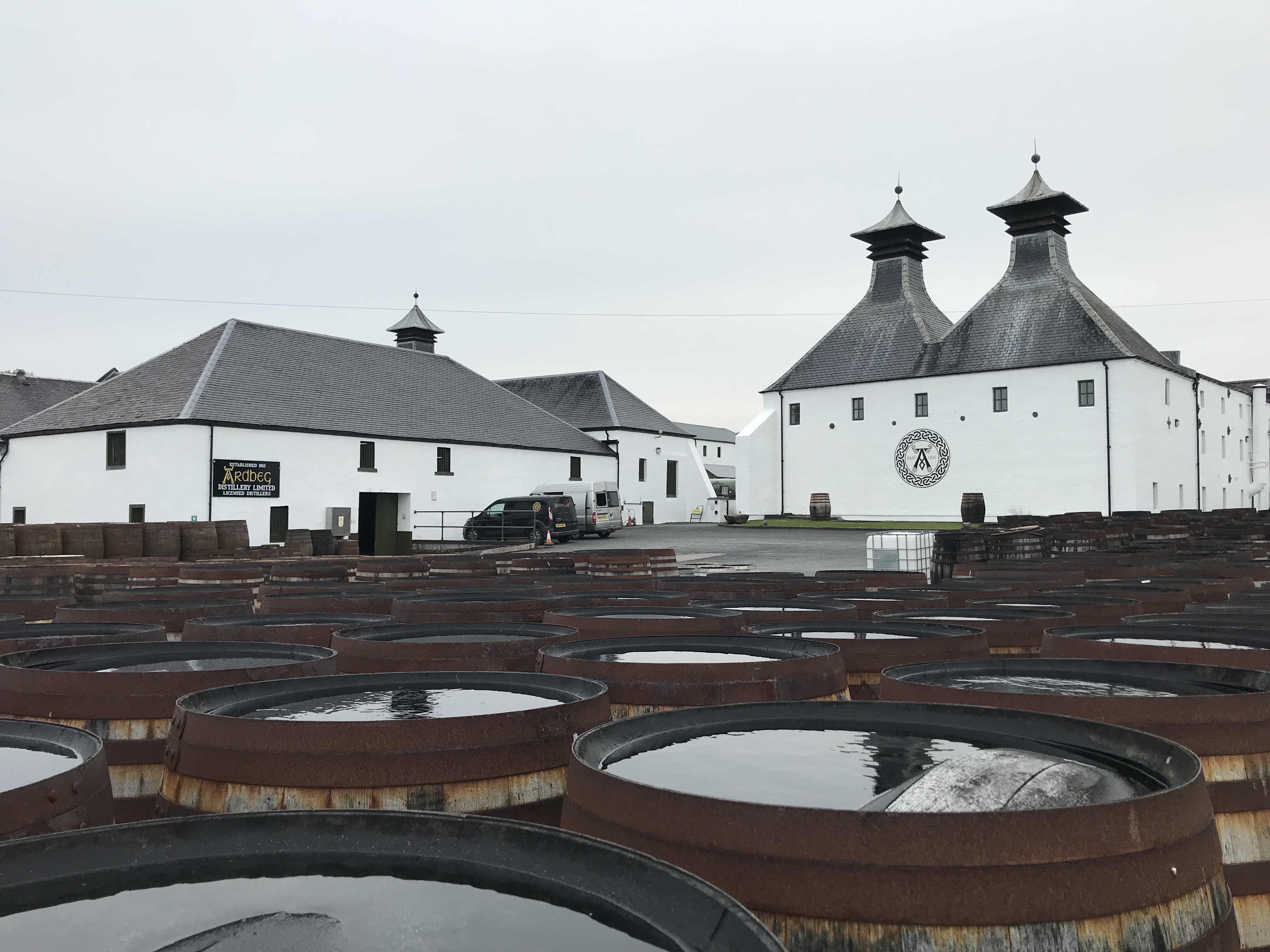View of white distillery buildings with slate grey roofing. A large collection of barrels is present in the foreground.