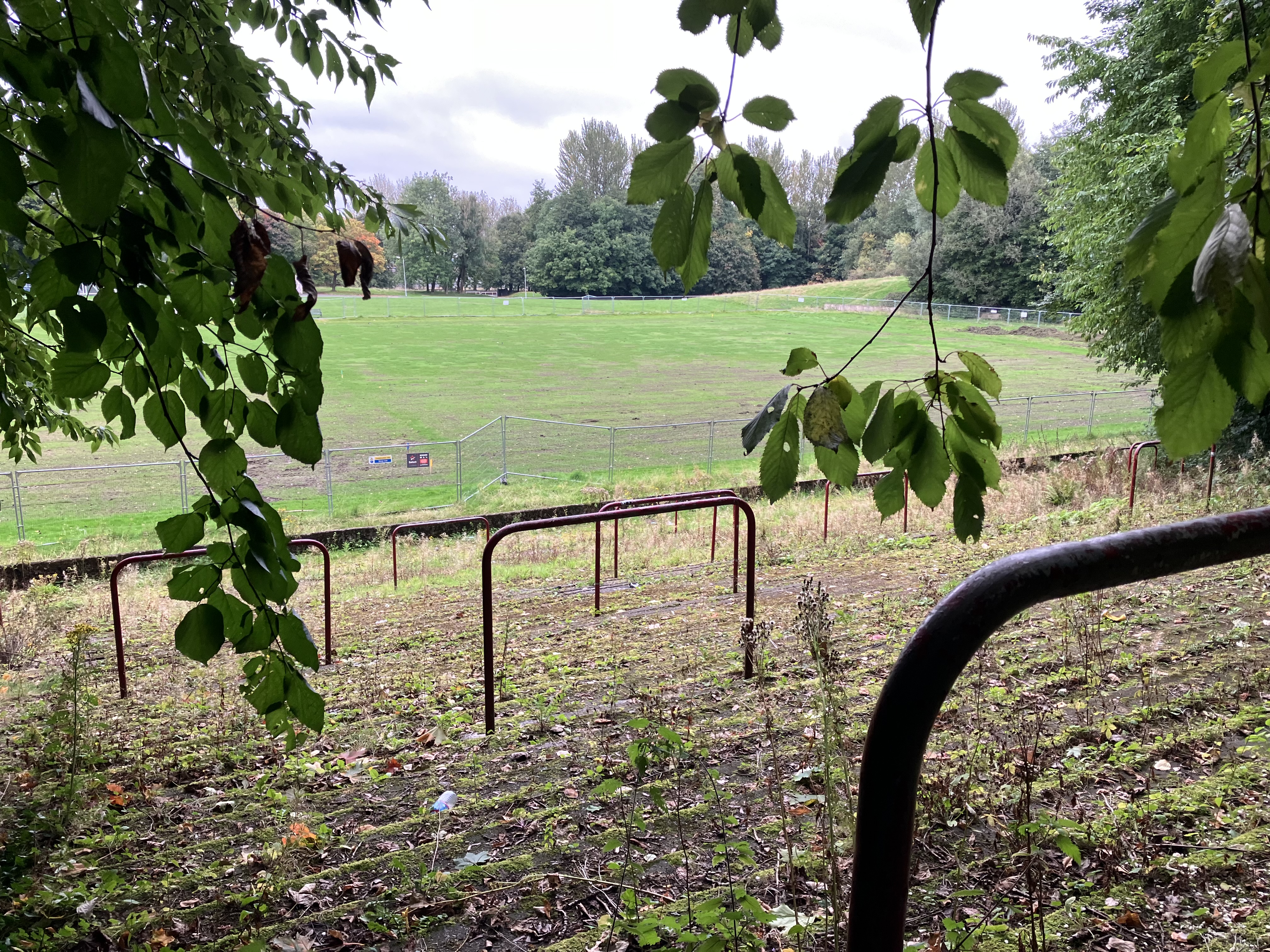 New Cathkin Park, former football Ground. View from the terrace steps across football pitch with trees in the foreground, the terrace steps are covered in moss and leaves with nettles growing throughout. Metal fencing surrounds the football pitch. Taken on a cloudy day. 