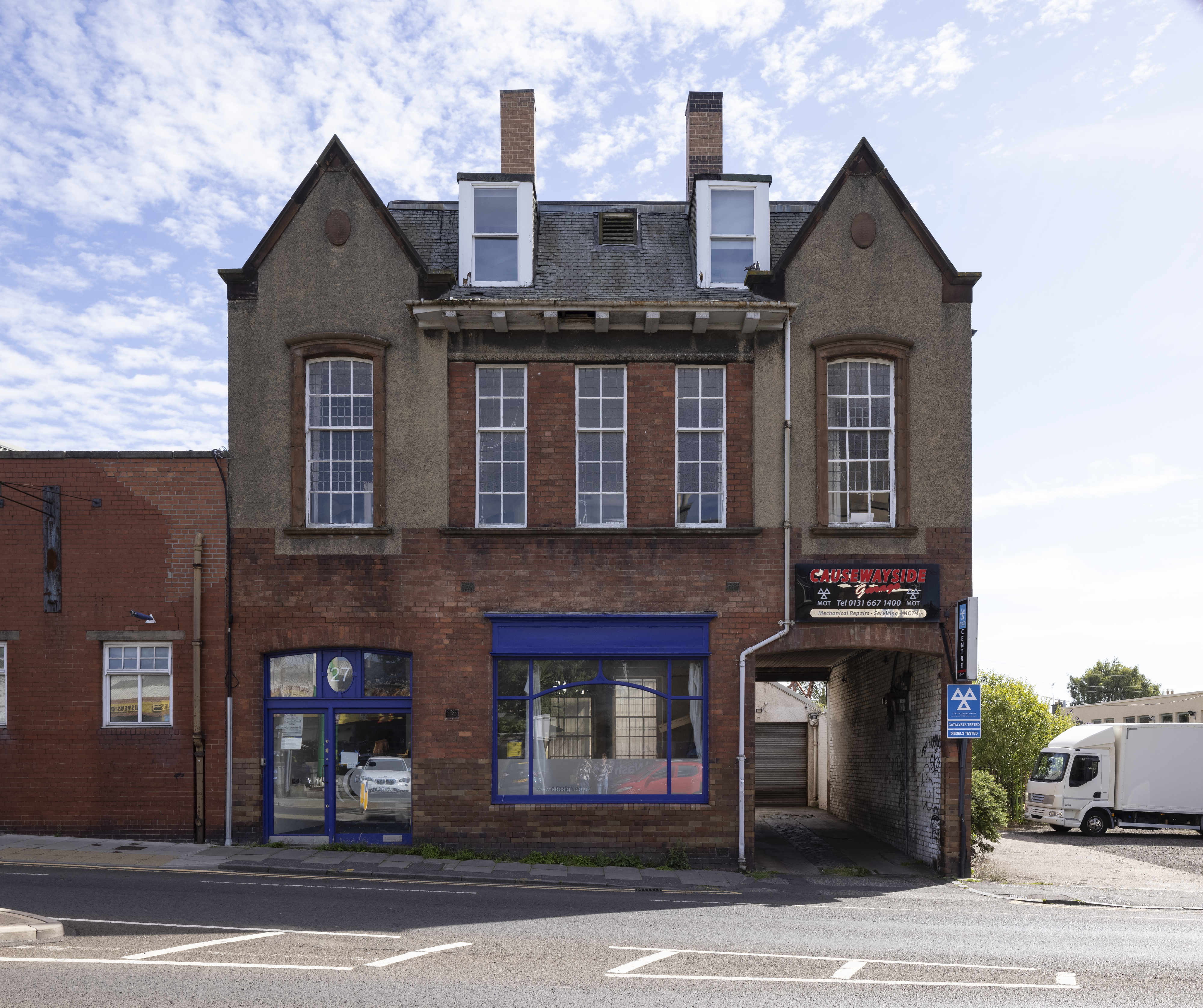 A multi‑storey brick building, formerly the Lads’ Institute, with tall first‑floor windows, dormer windows in the roof, and blue‑framed doors and windows at street level. A modern business sign reading “Causewayside” is mounted on the right side, and a narrow driveway leads to a rear garage. A white truck is parked beside the building towards the rear..