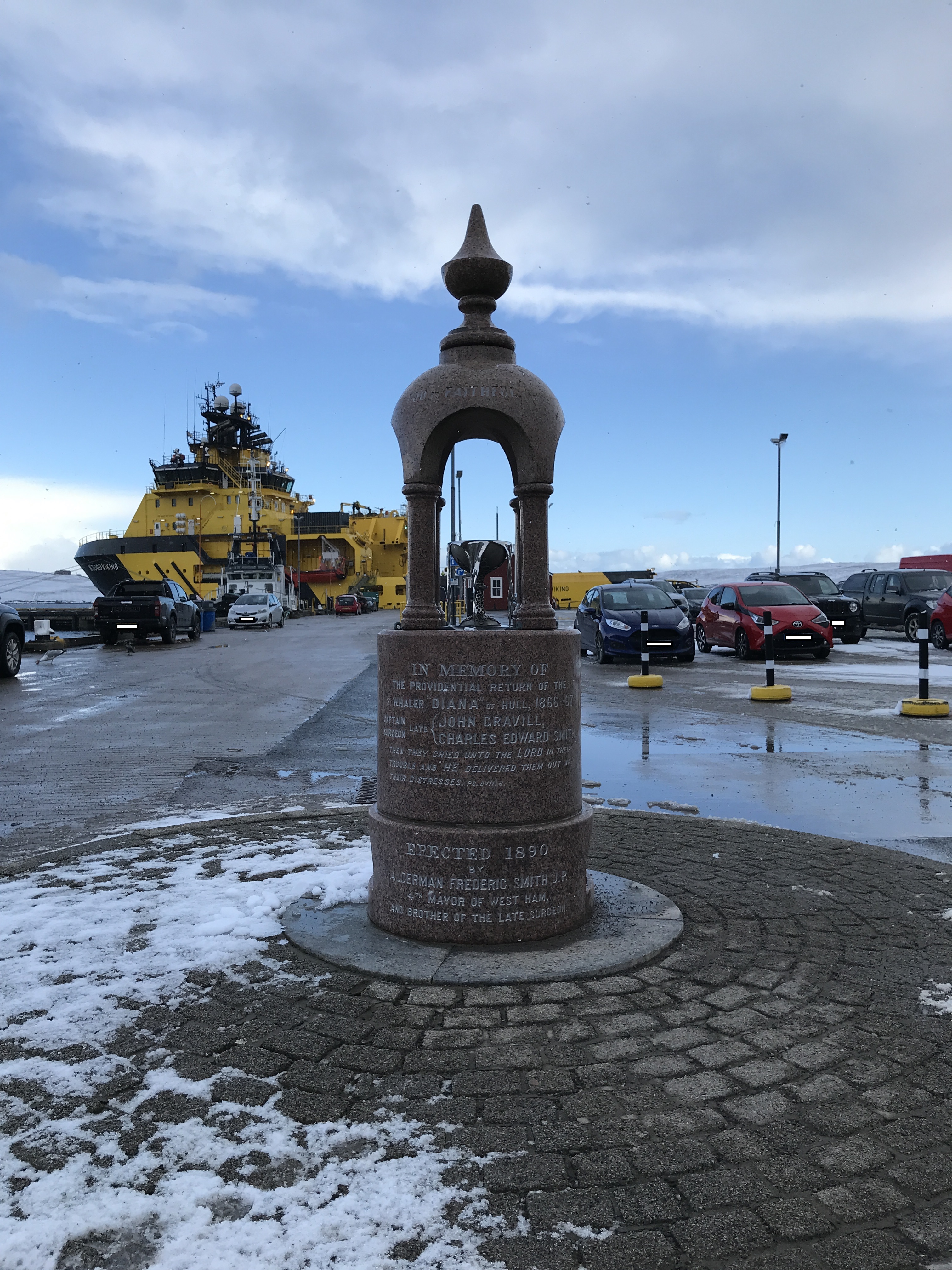 Memorial drinking fountain to the Diana. The fountain is pink and grey granite, and sits atop top blocks with lengthy 'In memory of' inscription. Snow covered the bricks stones surrounding the fountain. And in the background there is a carpark and a docked yellow ship.