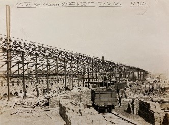 An industrial building under construction, with only metal frame erected. Bricks are stacked in neat piles, and a small rail cart sits empty on a track. 