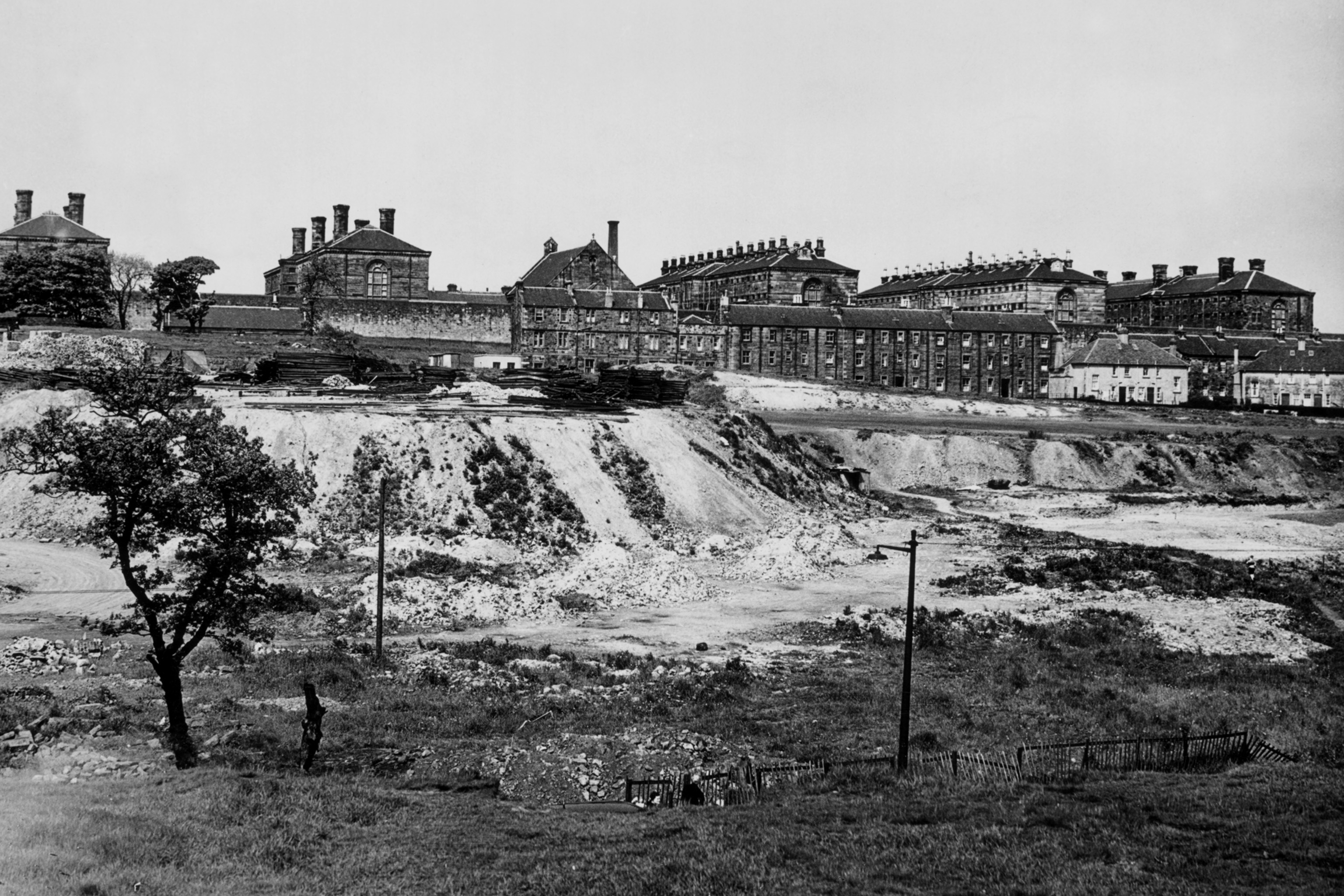 Landscape, black and white photo of Barlinnie prison from across a field. In front of the many prison buildings is a large bank that drops into the field.