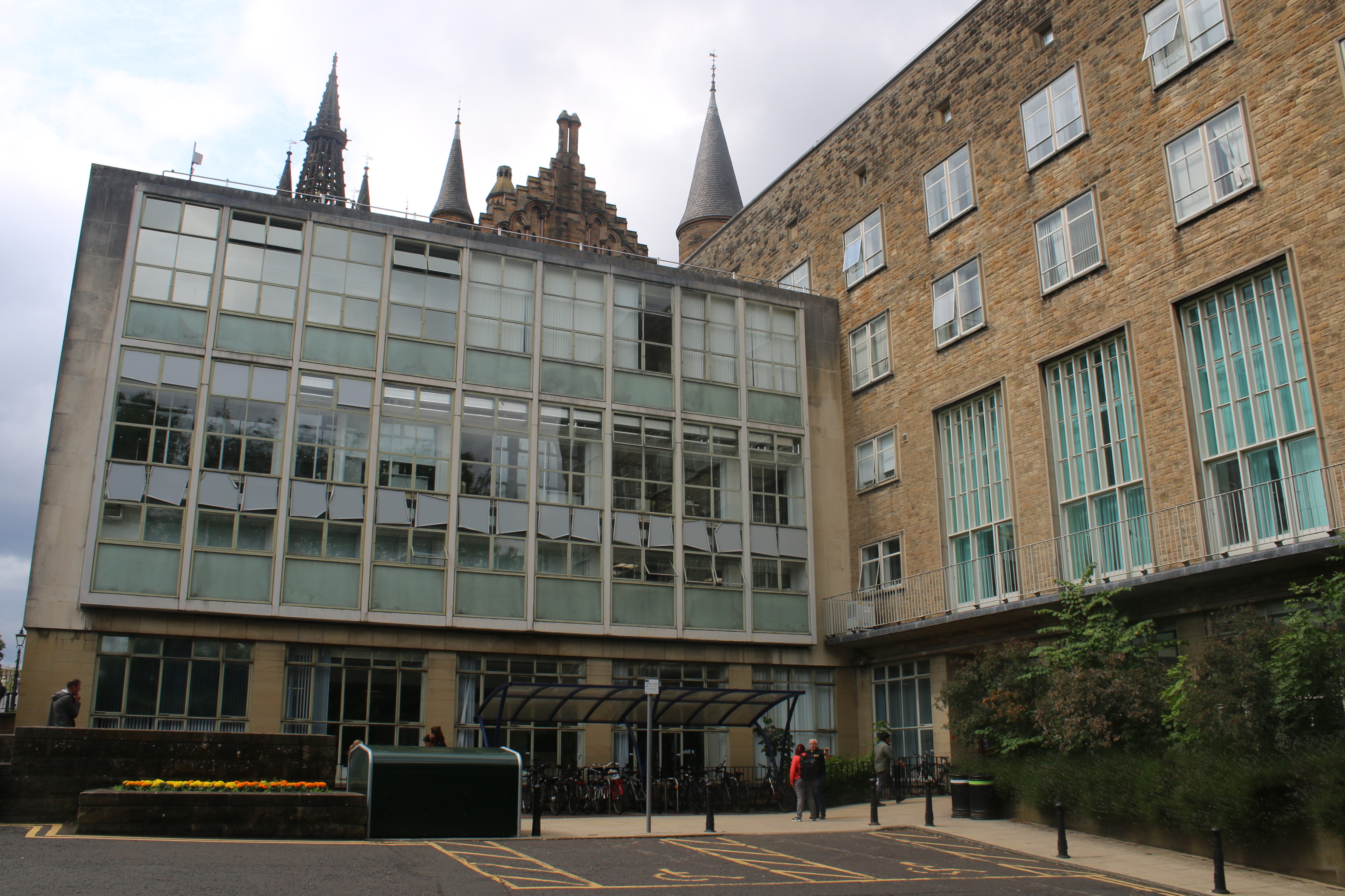 Main elevation of the James Watt Building with the Gilbert Scott Building in the background. Left side of building as facing photo is almost entirely windows, while the right side facing is a mixture of yellow bricks with windows are regular spacing. In front of the entrance is two disabled parking spaces and a bike rack. It appears to be a gloomy day.