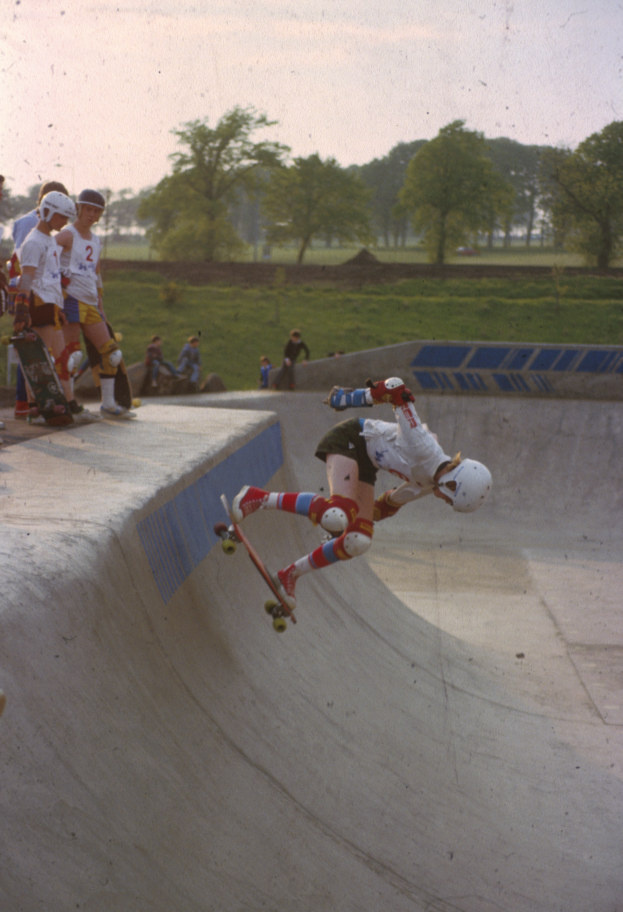 Archive image of skate-boarder with white top and helmet dropping into bowl, watched by skateboarders on ledge to left