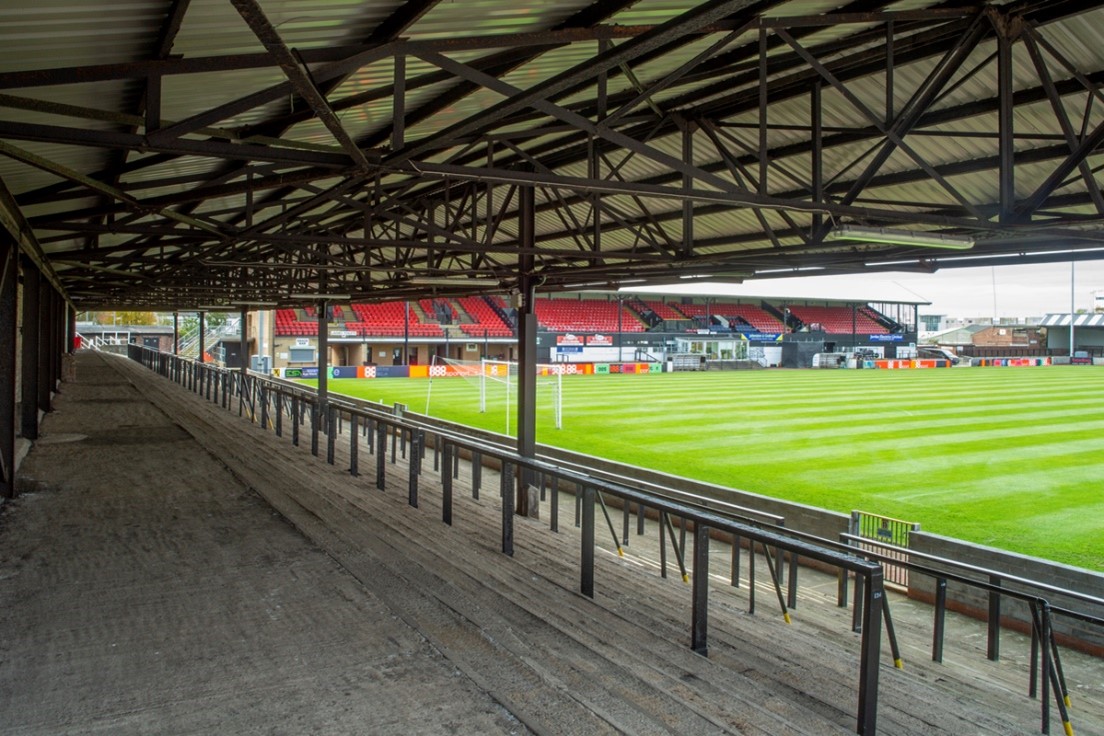 View from beneath a covered terrace inside a football ground, looking across a green pitch with goalposts, toward a stand with rows of red seats, supported by a steel roof structure and lined with railings along the empty concrete steps.