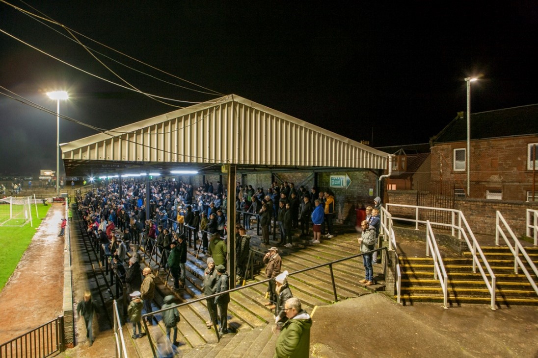 Covered football terrace at night filled with spectators standing on concrete steps, viewed from the side near the pitch, with bright floodlights illuminating the scene, a goalpost visible on the left, and nearby buildings and railings surrounding the stadium.