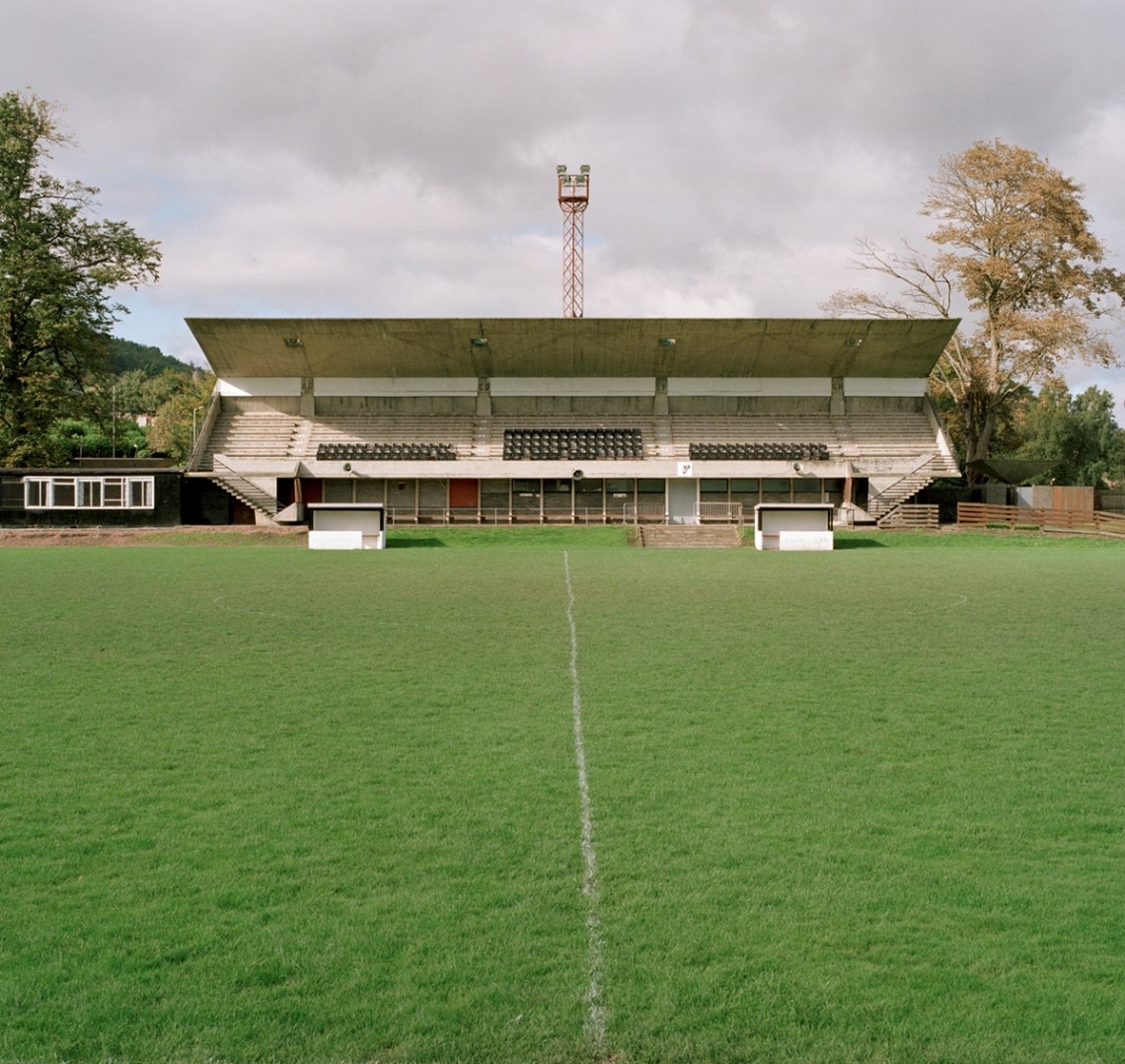 Modern concrete stand at Gala. The stand is empty from our central pitch view, marked by a white halfway line trailing away toward the concrete stand, with small white empty dugouts either side. There are rows of seats under a wide roof, flanked by staircases and trees, beneath a cloudy sky.
