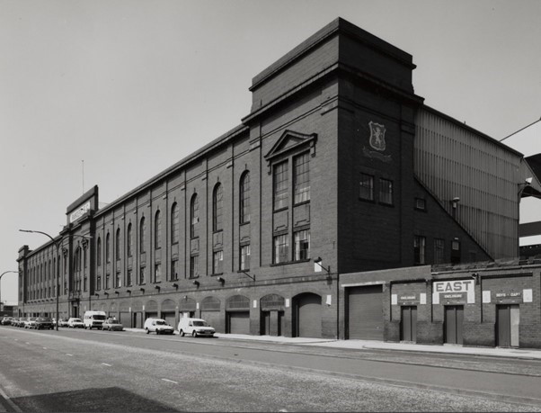 Black-and-white photograph of a large brick football stadium exterior along a street, featuring a long facade with arched windows and a taller central structure, with several parked vintage cars and entrances marked by signage including “EAST.”
