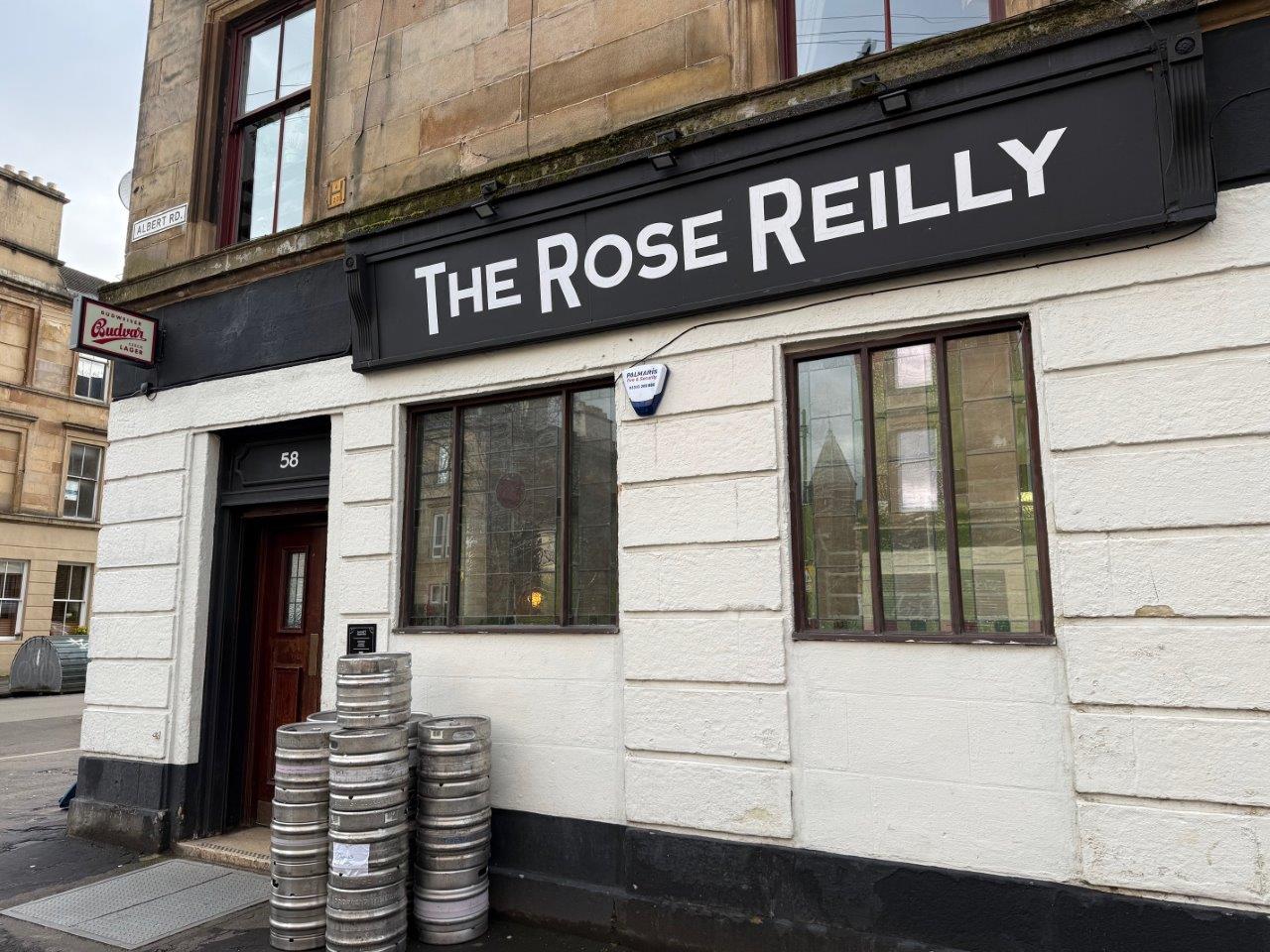 Exterior of a traditional corner pub with a black sign reading “The Rose Reilly” above white-painted walls, large windows, and a wooden door marked “58,” with several stacked metal kegs outside and a Budweiser sign mounted on the adjacent wall.